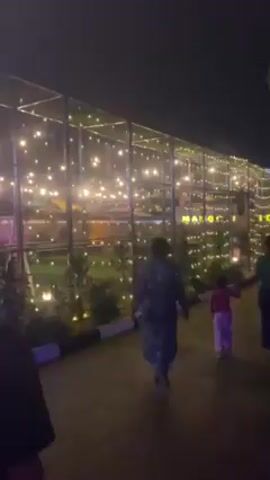 The video shows an outdoor scene at night, featuring a group of people walking towards a brightly lit building. The sky is dark, indicating evening or night time.

In the foreground, the back of a person wearing a dark t-shirt is visible, walking away from the camera. Ahead of this person, several other individuals are walking in the same direction. These include an adult wearing a patterned blue and white garment, holding hands with a small child in pink pants and a purple top. Further ahead, another adult wearing dark clothing is also walking.

To the right, there's a large structure, possibly a restaurant or event space, that is heavily decorated with numerous string lights or fairy lights. These lights are bright and warm-toned, creating a festive and inviting atmosphere. The structure appears to be made of glass or transparent panels, allowing the interior lights and decorations to be seen. Inside the structure, more lights and some green foliage are visible. The word "MANGI" or "MANGS" is spelled out in large, illuminated yellow letters on the side of this building.

Along the base of the lit structure, there's a low white and black striped curb, separating the walking path from the building. To the far right, past the main lit structure, another large sign is partially visible, which seems to include the words "HAPPY" and "NEW YEAR," suggesting a celebratory context, possibly around a holiday. There are also trees and darker foliage visible on the right side of the frame, contrasting with the bright lights.

The overall impression is that of people arriving at or walking through a lively, decorated outdoor venue or commercial space during the evening.