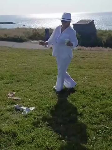 A woman in white clothing and a white hat dances in a grassy field near the ocean. There are sandals on the ground near her. In the background, there is a small building and the sea. The video then transitions to show another woman in a straw hat and jeans, also in a grassy area with the sea and some ruins in the background.