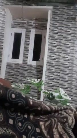 The video shows a close-up of a person's feet and legs, covered by a patterned cloth, as they are resting. The background is a residential building with gray tiled walls and white trim. There are two windows visible on the building, and a white door is also seen further down. Green plants are in front of the building. The scene appears to be outdoors, possibly in a yard or porch area of a home. The lighting suggests it might be evening or nighttime. The overall atmosphere is relaxed and casual. No text is visible in the video.