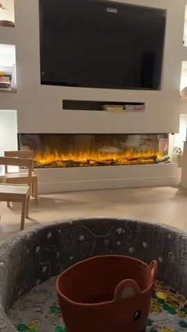 The video shows a close-up view of a modern electric fireplace with glowing logs. The flames are a vibrant mix of orange, yellow, and purple, creating a warm and inviting atmosphere. The fireplace is set into a white mantelpiece, and the reflection of the flames can be seen on the glass front. In the foreground, a soft, grey dog bed with white star patterns is visible, suggesting a cozy living space. The overall impression is one of comfort and relaxation, with the fireplace serving as a focal point in the room.