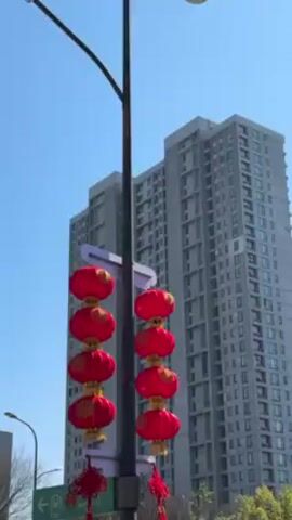 The video shows a street in an urban area.  Two strings of four red lanterns hang from a tall, gray light pole. The lanterns have golden accents. In the background is a tall, gray apartment building under a clear, blue sky. A car passes by on the road. A street sign with arrows is visible.  Some trees are partially visible.