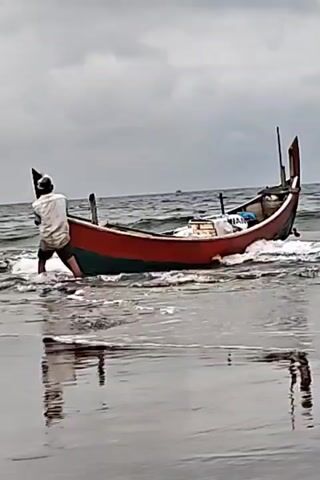 A person stands in shallow water, pushing against the side of a red and green boat. The boat is tilted, with its bow pointing upwards and slightly out of the water. The person is facing away from the camera, wearing a white shirt and dark shorts. The boat appears to be on a beach, with waves gently lapping at its hull. The water reflects the sky and the boat. The sky is overcast and the sea is choppy.