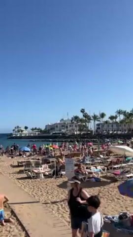 Here's a forensic description of the video:

The video depicts a crowded beach on a sunny day, likely a popular tourist destination. The sky is clear and blue, indicating fair weather. The beach is sandy and densely populated with people of various ages and ethnicities. Many individuals are sunbathing on lounge chairs or directly on the sand, while others are walking along the beach or gathered near the water's edge.

Numerous colorful beach umbrellas are scattered throughout the scene, providing shade. The ocean is visible in the background, with a rocky breakwater extending into the water.  Beyond the beach, there is a structure with a white facade, possibly a hotel or resort. Palm trees are also visible near this building. The camera appears to be handheld, with slight movements and adjustments in angle.