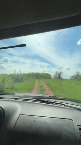 The view from inside a car shows a dirt road on a sunny day. The sky is blue with scattered white clouds. Green vegetation borders the road. The car's dashboard is visible in the foreground, and the windshield is slightly dirty. The wipers intermittently move across the glass.