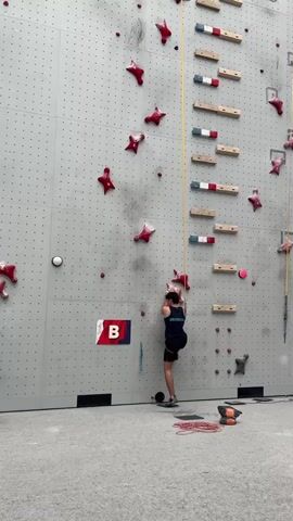 A person is rock climbing indoors on a tall, grey wall with numerous red and pink star-shaped climbing holds, as well as wooden horizontal rungs. The climber, wearing a dark tank top and shorts, is ascending the wall, reaching for a hold. A large rectangular banner with the letters "B" in white on a red and blue background is affixed to the wall. Red climbing ropes are coiled on the floor at the base of the wall, along with other climbing gear. The lighting suggests an indoor environment, likely during the day. The scene is focused on the athletic activity of climbing. Text visible includes "GRAFFITI" on the climber's tank top and the letter "B" on the banner.