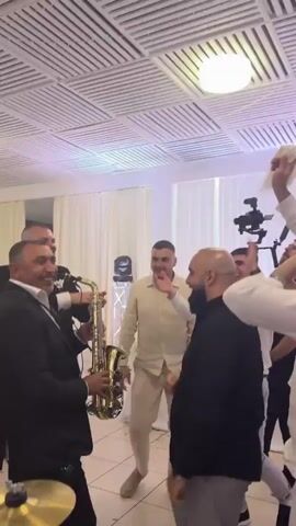 Several men are dancing at what appears to be a wedding reception. One man is playing the saxophone. The men dancing are mostly dressed in lighter colors, white and beige, while one is wearing a black shirt. Some are waving white cloths in the air. A video camera is set up behind them. The room has white walls, a drop ceiling, and a large white curtain behind the group. The room is lit with blue and white light.