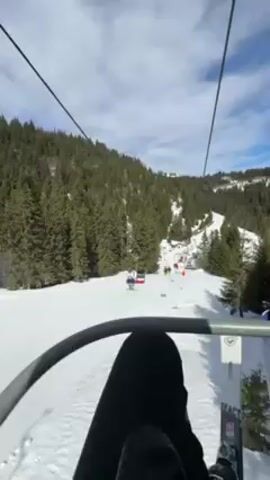 A first-person point of view shot taken on a ski lift moving uphill on a snowy mountain. The skier is wearing black pants and has their skis attached to their boots. The skis are white with black and blue markings. In the background, a ski run is visible with several skiers on it, and the surrounding landscape is covered in evergreen trees. The sky is blue with scattered white clouds. The ski lift chair is metal and provides a safety bar in front of the skier.
