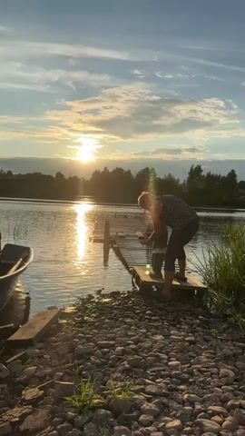A woman with blonde hair, wearing a striped top and dark pants, stands on a wooden dock at the edge of a body of water. She is holding a small child, also in dark clothing, and appears to be interacting with them near the water's edge. The sun is setting, casting a warm glow over the scene, with a boat visible to the left and trees lining the opposite shore. The foreground is covered in pebbles and green grass. The woman gestures towards the water, possibly indicating something to the child.