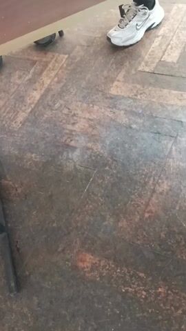 The video shows a worn wooden floor in a herringbone pattern. In the top right corner of the frame, there is a white sneaker with black accents. To the left, a corner of a wooden table is visible. There are sounds of a crowd and what seems to be shouting or excited sounds.
