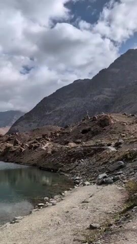 A static shot pans across a serene mountainous landscape. In the foreground, a tranquil lake reflects the cloudy sky and the rugged, rocky terrain that surrounds it. Steep, brown mountains dominate the background, their peaks partially obscured by dramatic, white clouds. The path leading alongside the lake suggests a popular hiking or tourist destination.