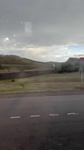 This media file captures a view from a vehicle traveling along a road, likely in a rural or semi-rural setting in Qarqaraly, Kazakhstan. The scene is dominated by a vast, open landscape with rolling hills in the background under a cloudy sky.

The primary subjects are man-made structures related to transportation and commerce. Prominently featured is a gas station, branded "ASIA Oil," with a distinctive red and white canopy. Adjacent to it is a gas price display sign, showing prices in a foreign currency, though the specific numerals are somewhat blurred. Further down the road, other buildings are visible, including what appears to be a small cluster of shops or roadside establishments.

The activity taking place is the movement of vehicles. An orange dump truck is driving on the road towards the viewer, indicating it's traveling in the opposite direction of the vehicle the footage is being filmed from. There are no people clearly visible in the frame, suggesting the focus is on the journey and the surroundings rather than human interaction.

Notable details include the weather, which appears overcast with scattered patches of sunlight breaking through the clouds, suggesting a dynamic or changeable sky. The road surface appears wet, indicating recent rain, which also causes reflections and streaks on the window through which the footage is taken, adding a slight blur and obscuring some details. The time of day is difficult to determine precisely due to the cloud cover, but the lighting suggests it is daytime, possibly late afternoon or early evening. The landscape is characterized by dry vegetation and sparse trees, consistent with a steppe or semi-arid environment.

The visible text includes the brand "ASIA Oil" on the gas station and the prices on the gas pump display. The overall impression is one of a journey through a functional yet somewhat desolate landscape, common in transit routes in less densely populated regions.