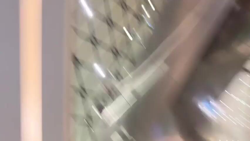 A close-up, upward-facing video shows the top of a person's head with dark hair pulled into a bun, wearing a black clip or accessory. In the background, a rack of shoes hangs from the wall. To the right, a vertical light fixture illuminates a decorative ceiling pattern. The lighting is soft, and the overall scene appears to be indoors, possibly a retail environment.