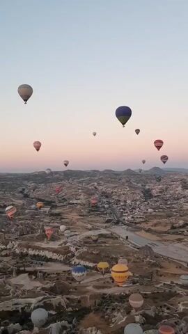 The video shows a panoramic aerial view of a landscape dotted with numerous hot air balloons. The balloons are of various colors and sizes and are scattered across the sky. The landscape below appears to be a rugged, mountainous terrain with some signs of human habitation, possibly a town or village. The sky is a gradient of soft pinks and blues, suggesting either sunrise or sunset. The camera is panning slowly across the scene, capturing the full scope of the hot air balloon spectacle.