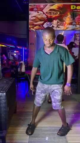 A sweaty man is dancing, moving with the music, in front of what appears to be a bar or restaurant. He's wearing a dark green polo shirt, camo shorts, and black sandals. He's holding a small can in one hand. Other people are visible in the background, some standing near an open doorway, others seated. The lighting is dim and colorful, with neon lights and a flashing light. There's a large speaker next to the man. A sign with food pictures is displayed on the building.