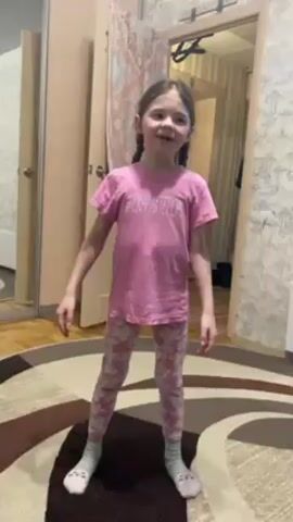 A young girl stands on a circular rug in a room and dances. She has brown hair in pigtails and wears a pink t-shirt, pink and white leggings, and blue socks. She makes various gestures with her hands as she dances. The room contains a mirror, a wooden door, and patterned wallpaper. The rug has brown and white patterns.
