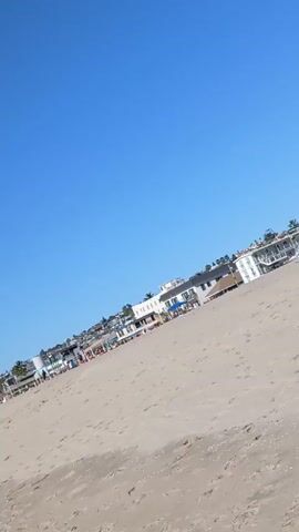 The video shows a beach on a sunny day. The sand is light brown, and there are several people enjoying the weather. Some are lying on towels, while others are sitting in beach chairs. There are also some palm trees scattered along the beach. In the background, there are several buildings, including houses and hotels. The sky is clear and blue. The camera pans across the scene, showing different groups of people and parts of the beach.