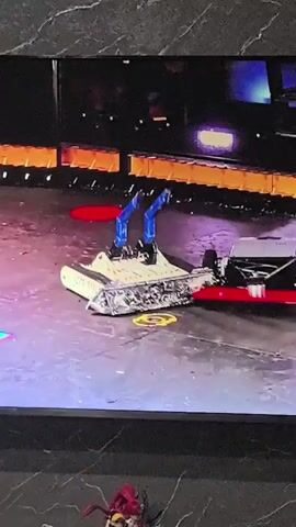 The video shows a robot combat competition. Two robots are facing each other in an arena. The robot on the left is white and tank-like, with two blue arms extended upwards. The robot on the right is black, with wheels and a flat, angled surface on top. The robots move towards each other and collide. Sparks are visible during the collision. The arena floor is a mottled grey with colorful tiles.
