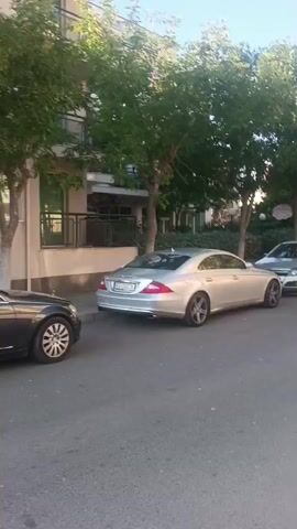 This media file shows a street scene in Nesebar, Bulgaria, likely in the late afternoon or early evening due to the warm, slanted sunlight and the long shadows. The weather appears to be clear and pleasant. The scene is a residential street lined with trees and parked cars.

The primary objects in the foreground are several parked cars, including a silver Mercedes-Benz CLS, a silver hatchback (possibly an Opel Astra), a black convertible (possibly an Audi TT), and a red hatchback. Further down the street, more cars are visible, suggesting a densely populated area or a popular parking spot.

In the background, a multi-story building with balconies and windows is visible, partially obscured by trees. The building appears to be an apartment complex or hotel. The trees are mature and leafy, providing shade.

The activity taking place is passive observation of the streetscape. There are no people prominently featured in the initial frames, but a man wearing a white t-shirt and blue shorts walks away from the camera on the sidewalk in the later frames. His posture and attire suggest a relaxed, leisurely walk on a warm day. The overall mood is calm and ordinary, depicting a typical urban or semi-urban environment.

Visible text includes license plates on the cars. The silver Mercedes has a license plate that appears to read "PB 0208 SB". The silver hatchback has a license plate that starts with "SL". The black convertible's license plate is partially visible and seems to start with "SL". The red hatchback's license plate is illegible. The overall setting is a paved street with a sidewalk made of interlocking pavers. The architecture of the building and the style of the cars suggest a European location.