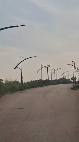 The video shows a paved road curving to the right and ascending slightly. The sky is overcast, with the sun visible through the haze. On either side of the road, there are tall lamp posts with curved arms, many of which are illuminated. There are also utility poles with electrical wires visible in the distance. The road is bordered by a low wall or embankment on the left, with trees and vegetation beyond it. On the right, there is a paved shoulder with some vegetation and a small signpost. The scene appears to be a rural or suburban area.