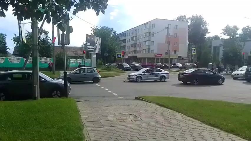 This media file captures a scene at a crossroad in Rechytsa, Belarus. The time of day appears to be daytime, under cloudy skies, suggesting overcast weather. Several vehicles are present, including a dark hatchback in the foreground on the left, a light-colored hatchback turning at the intersection, and a white car with markings that suggest it is a police vehicle, also maneuvering through the intersection. Other cars are visible in the background, some parked and others in motion.

The scene is set in a built-up area, with a multi-story apartment building dominating the background. Adjacent to it, a store with prominent signage can be seen. Trees line the sides of the road, and a sidewalk with brick paving is visible in the foreground. Traffic cones are placed on the road, indicating potential roadwork or a temporary traffic diversion. A traffic light is visible, showing a green signal for one direction of traffic.

The activity appears to be normal traffic flow, with vehicles navigating the intersection. The police car's presence might suggest an ongoing situation, but no overt signs of distress or emergency are apparent. The focus is on the vehicular movement within the intersection.

There is visible text on a billboard in the background, which is partially obscured and illegible. The police car has text on its side, but it is also too small and blurry to read clearly. The overall impression is a routine urban scene with moderate traffic activity.
