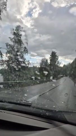 The video shows a drive on a wet road with rain on the windshield. The road is surrounded by trees and greenery, with a body of water visible to the left. The sky is cloudy with patches of blue sky. Several cars are seen on the road, including a red car that passes the viewer's car. The overall mood is somewhat dreary due to the weather, but the presence of nature and other vehicles suggests a normal day on a rural road. The time of day appears to be daytime, given the visible sky and lighting conditions. The location is identified as Novovilgovskoye Sel'skoye Poseleniye, Russia. There is no visible text in the video.