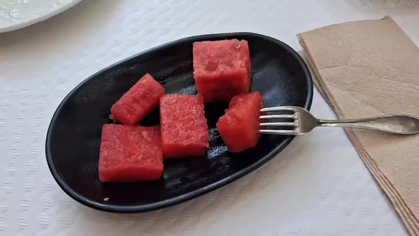 The media file shows a close-up shot of a black oval plate filled with several cubed pieces of watermelon. A silver fork is shown piercing one of the watermelon cubes, as if about to be eaten. The watermelon pieces are a vibrant red color. The plate rests on a textured white surface, possibly a tablecloth, with a light brown napkin placed to the right of the plate. The scene is well-lit, suggesting it is daytime. The focus is entirely on the food and the action of picking it up with the fork. There is no visible text in the image. The setting appears to be a casual dining or snack situation. Given the location context of Lloret de Mar, Spain, this could be a refreshing snack enjoyed during a vacation. The image evokes a sense of simple pleasure and enjoyment of fresh fruit.