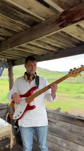 A man wearing a white shirt and blue jeans stands under a wooden canopy, playing a red bass guitar. He has headphones around his neck. The background shows a green, grassy landscape with a wooden fence. The camera is positioned at a medium shot, focusing on the man and his guitar. The lighting appears to be natural daylight.