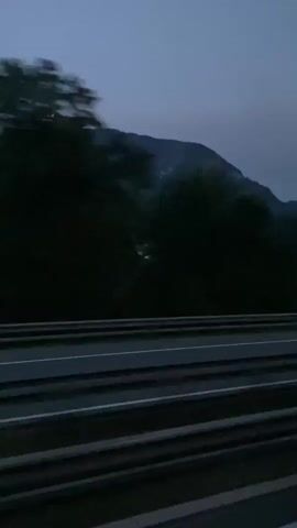 The video captures a side-view perspective from a moving vehicle, likely on a highway, as it travels alongside a dark, tree-lined landscape. In the distance, a large, silhouetted mountain range dominates the horizon under a twilight sky. The foreground shows multiple lanes of a road with white lines, separated by guardrails. The trees are densely packed and dark, obscuring much of the terrain behind them. As the vehicle moves, the trees and a distant structure blur, indicating speed. The overall atmosphere is dim and serene, suggesting travel at dusk or dawn.