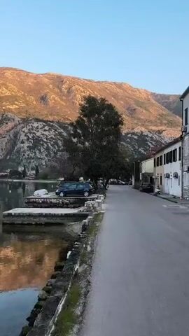 The video presents a static, daytime view of a coastal scene featuring a calm body of water, a road, and a backdrop of mountainous terrain. On the left, the water's surface reflects the colors of the sky and the mountains, with a stone pier extending into the water. A dark blue car is parked on the pier. Along the right side of the frame, a narrow paved road runs parallel to the water. Adjacent to the road are a few buildings, painted in light colors. A single tree with dense foliage stands between the road and the pier. In the background, a large mountain range rises steeply, its surface marked by rocky outcroppings and sparse vegetation. The sky is clear and light blue, suggesting either early morning or late afternoon light.