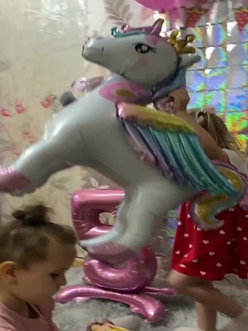 A birthday party is being celebrated in a room. Two young girls are present. The older girl, wearing a red dress with white hearts and a white top, is interacting with a large, inflatable unicorn. The younger girl, wearing a pink top, is sitting on the floor. A large pink balloon shaped like the number five is visible. The walls are decorated with a floral wallpaper and a shimmering backdrop.