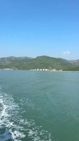 The video captures a view from a boat moving through calm, green water under a clear blue sky. In the distance, a lush green coastline dotted with buildings and topped with rolling hills stretches across the horizon. The water behind the boat shows the white wake of its passage.