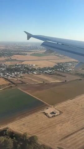 The aerial view shows an airplane wing on the right side of the frame, extending into the blue sky. Below, a rural landscape unfolds, characterized by patchwork fields in various shades of brown, green, and yellow, indicating agricultural activity. A small cluster of buildings, likely a village or settlement, is visible amidst the fields, with some structures appearing to be houses and others possibly industrial or agricultural facilities. In the distance, a city skyline with high-rise buildings can be seen on the horizon.

The time of day appears to be morning or late afternoon, given the clear blue sky and the direction of the sunlight, which casts shadows across the land. The weather seems fair and clear, conducive for flying. The landscape suggests a location with arid or semi-arid climate, with dry vegetation in some areas and cultivated fields in others. The presence of an airplane suggests a journey is in progress, possibly during takeoff or landing, given the low altitude. The scene evokes a sense of travel and exploration, offering a bird's-eye perspective of the earth. No visible text is present in the media. The location context provided is Kfar Truman, Israel, which aligns with the visual cues of a Mediterranean landscape.