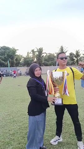 This media file features two people, a woman and a man, posing for a photo with a large golden trophy. The woman, on the left, is wearing a black hijab and a black top with long sleeves, paired with light blue denim jeans. She is holding the trophy with both hands, looking towards the camera with a slight smile. The man, on the right, is wearing a bright yellow t-shirt and black pants, with sunglasses on. He is also holding the trophy and has his right arm raised in a celebratory pose, with his fist clenched. The trophy is decorated with colorful ribbons.

They are standing on a grassy field, likely a sports ground, with a blurred background of trees, buildings, and other people in the distance. The sky is overcast, suggesting it might be late afternoon or early evening. The overall mood appears to be one of celebration and achievement, possibly at a sporting event or competition. The text on the trophy is partially visible, suggesting it is a prize for a winner. The setting is outdoors and appears to be in a community or public space given the presence of other individuals and what looks like a fence or boundary in the background. The lighting is soft, consistent with daylight, but without direct sunlight.