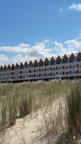 The video opens with a view of a long, multi-story building with a distinctive pointed roofline, set against a bright blue sky with scattered white clouds. In the foreground, tall, dry grass grows in sandy dunes. The camera then pans to the right, revealing more of the sandy landscape, with the building receding into the background. Wooden posts are visible in the sand, one with a small plant growing near it. The video continues to pan right, showing a wide expanse of dunes and a distant coastline under a clear blue sky. The scene appears to be a coastal resort or town with a beach and dune system. There are no people or animals visible in the video. The overall impression is one of a sunny, serene beach environment.
