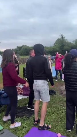 A group of people are gathered outdoors in what appears to be a park or campsite. The sky is overcast and the lighting is dim, suggesting it is either early morning or late afternoon. There are trees in the background, and some tents or shelters are visible. Several people are standing and talking, while others appear to be exercising or dancing. One person is wearing a black puffer vest and a blue baseball cap, facing away from the camera. Another person in the foreground is wearing a black hoodie and denim shorts. A pink yoga mat is visible on the ground. A portable speaker is also present. Some people are wearing casual clothing like hoodies, t-shirts, and shorts, while others are dressed in puffer vests and jackets, indicating cooler weather. The overall atmosphere seems relaxed and social, with a hint of an organized outdoor activity or gathering.
