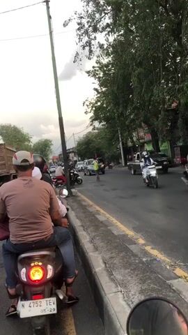 A street scene with multiple motorcycles and some cars. In the foreground, a person wearing a light brown shirt and a white baseball cap is sitting on a scooter, facing away from the camera. The scooter has a license plate that reads "N 4752 TEV 05-29". Another person is sitting behind the driver. To the right, another motorcycle is partially visible in the frame with a rider wearing a blue jacket and a white helmet. Further down the street, there are several other motorcycles and cars, including a silver SUV and a black pickup truck. One person in a yellow vest appears to be directing traffic. The scene is outdoors with trees lining the right side of the road and a cloudy sky.