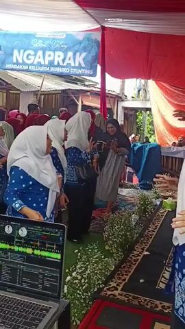 The video shows a group of women dancing at an outdoor event, likely a community gathering in Indonesia, judging by the language on the banner.  The women, dressed in blue patterned tops and hijabs, are clapping and moving to music. A laptop with audio mixing software sits on a table in the foreground, suggesting the event is professionally supported. The setting includes a tent-like structure with red and white fabric, decorated with flowers. There are other people milling around, some filming with their phones.  The event seems to be related to raising awareness about stunting, as indicated by the text on the banner.
