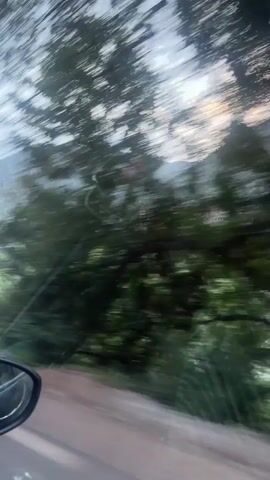 The video captures a scenic drive through a mountainous region in Taljarft, Morocco, viewed from the perspective of a passenger inside a car. The car is moving along an unpaved, dirt road, with the side mirror visible in the bottom left corner.

The landscape features rugged mountains covered in green vegetation, including numerous trees and shrubs, particularly in the foreground and lower slopes. A small, traditional village is nestled into the side of a distant mountain, characterized by buildings that appear to be made of earth or reddish-brown materials, with a prominent structure (possibly a mosque or a central building with a tower) standing out.

The sky is overcast, suggesting a cloudy day or a time of day with diffused light, possibly early morning or late afternoon. As the vehicle proceeds, a telephone pole briefly comes into view, followed by another car passing in the opposite direction. A pile of blue barrels is visible on the roadside at one point. The overall setting is rural and mountainous.