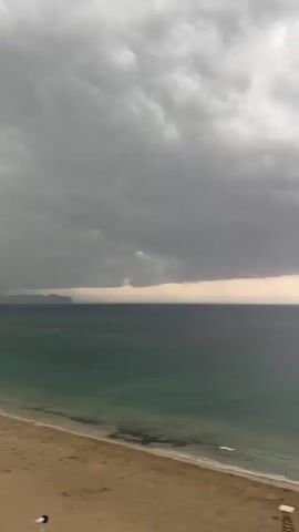 The video shows a view of a beach and the sea under a cloudy sky. In the foreground, there is a sandy beach with some shadows and a lifeguard chair. The sea is a greenish-blue color and there are small waves breaking on the shore. In the background, there is a large mountain on the right side of the frame and a distant landmass on the left. The sky is filled with dark, heavy clouds, suggesting an approaching storm. There are no people clearly visible in the video except for one person in the far distance on the beach in the fourth frame. There is a small boat in the water in the fourth frame. The overall atmosphere is one of impending weather change.
