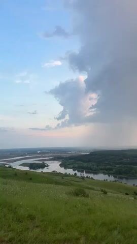 The video shows a landscape with a river and trees. In the foreground, there is a grassy hill with some wildflowers. In the background, there is a river with a few islands and a forest. The sky is cloudy with some patches of sunlight. The video progresses to reveal a ruined brick structure, resembling an archway, in the foreground. The archway is made of weathered red bricks. The video shows the scene through the archway, with the river and forest visible beyond. The sky continues to be cloudy, with a sunset glow appearing.