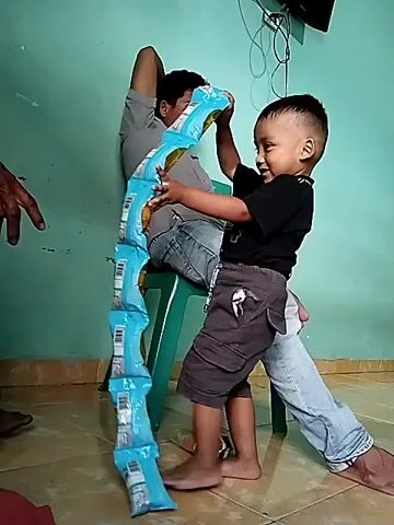 A man is sitting on a green plastic chair with his legs outstretched. A young boy is standing in front of him, holding a long string of blue and yellow snack packages. The boy is attaching the packages to the man's legs. The man is wearing a grey t-shirt and blue jeans. The boy is wearing a black t-shirt and grey shorts. The background is a light blue wall with a window or shelf visible at the top right. The floor is tiled. Another person's hand is visible on the left side of the frame, reaching towards the snack packages.