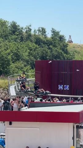 A group of people are on a roller coaster ride. The roller coaster is red and white and is on a track that is going up a hill. There are trees and a blue sky in the background. There are also some people standing in line to get on the roller coaster. The people on the roller coaster are wearing safety restraints and are smiling and laughing. The roller coaster is moving at a fast speed.