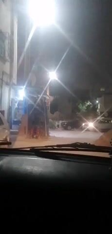 The video shows a night-time view from inside a vehicle, looking out through the windshield. The windshield wipers are visible in the foreground. In the background, a street is illuminated by a bright overhead light and the headlights of approaching cars. A white car with its headlights on is clearly visible in the middle ground, appearing to be moving towards the viewer. To the left of the street, there is a building with a storefront that is partially lit, revealing some shelves and items inside. The overall environment appears to be a street in a town or city at night. There are no people clearly visible within the frame. The atmosphere is somewhat hazy, possibly due to dust or moisture in the air.