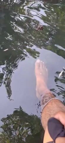 A person's bare foot and lower leg are submerged in murky water. The foot is visible, with toes clearly defined. The water is dark and somewhat opaque, with reflections of trees or foliage on the surface. The person's leg is hairy above the water line. The scene appears to be outdoors, possibly a stream or pond. The camera is angled downwards, focusing on the foot in the water.