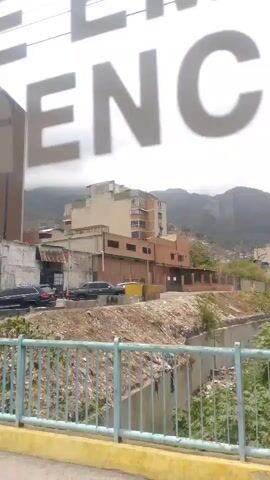 The camera films a city street from a moving vehicle. The street is lined with various businesses and buildings, including a liquor store called “Joselina”.  A tall building is visible in the background. People are walking on the sidewalks. Vehicles are parked and driving on the street. The sky is overcast. A banner with large words, “DE EMERGENCIA” is overlaid on the video.