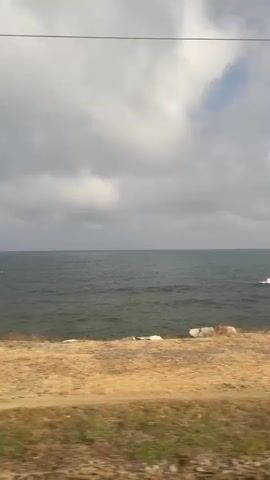 The video captures a view of the ocean from a moving vehicle. The sea is a dark blue-green with moderate waves and some whitecaps. The sky is overcast with large, fluffy grey clouds, but there are glimpses of blue sky and brighter clouds. The foreground consists of dry, yellowish-brown grass and some rocks along the coastline. There are also some green bushes and reeds visible, as well as a single pole with what appears to be a light fixture on top. The camera perspective suggests the footage was taken from a car or train traveling parallel to the coast.
