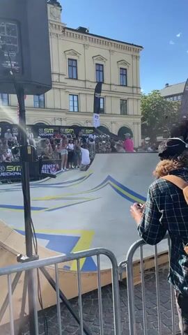This media file depicts a skateboarding event taking place outdoors on a sunny day, likely in Karlstad, Sweden, as suggested by the visible text. The scene is set in a public square or park area, with a large, ornate building serving as a backdrop. Spectators are gathered, seated on bleachers and standing around the perimeter, observing the competition.

The primary focus is on a skateboarder in a white t-shirt performing a trick on a ramp. The ramp is designed with distinct yellow and blue stripes. To the left, a large speaker on a stand is visible, suggesting this is a public event with amplified sound. A metal barrier separates the spectators from the action.

Several people are visible in the foreground and midground. The skateboarder is actively engaged in the sport. Spectators appear to be enjoying the event, with some holding phones to record or take pictures. The person in the immediate foreground, with their back to the camera, is wearing a plaid shirt and a backpack, also holding a phone.

Visible text includes "SM VECKAN" on banners and flags, indicating this is part of a Swedish championship event. Other text, partially visible, reads "SVENSKA" which likely refers to "Swedish." The weather appears to be fair and warm, with a clear blue sky. The overall atmosphere seems energetic and festive, typical of a sporting event.