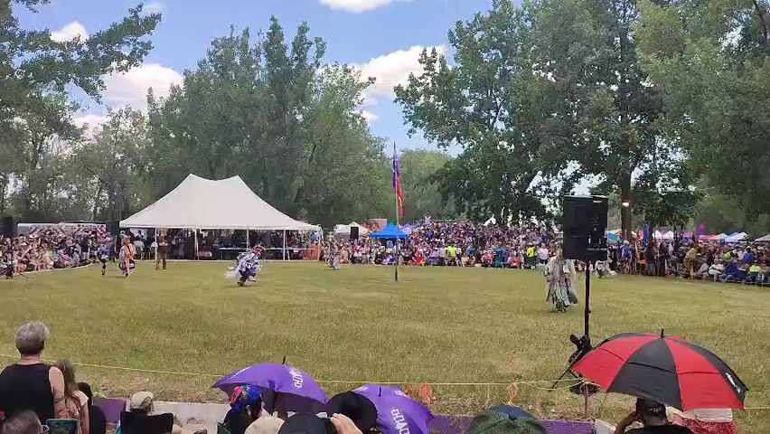 This video shows an outdoor event with a large crowd of people gathered on a grassy field. In the foreground, several individuals are seated and watching the event, with some holding umbrellas. In the middle ground, there are dancers in traditional Native American regalia performing on the grass. A large white tent is set up behind the dancers, and a blue tent is visible further back. A flagpole with a red and purple flag stands in the background. The area is surrounded by trees, and the sky is partly cloudy. The overall atmosphere appears festive and celebratory.