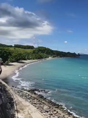 The video shows a scenic view of a tropical beach and ocean. The water is a clear turquoise color and the sky is blue with scattered clouds. There is a small beach with dark sand, and a rocky shoreline in the foreground. Green vegetation covers the hill in the background, where a few telecommunications towers are visible. The camera pans slowly to the right, revealing more of the ocean and the distant coastline.
