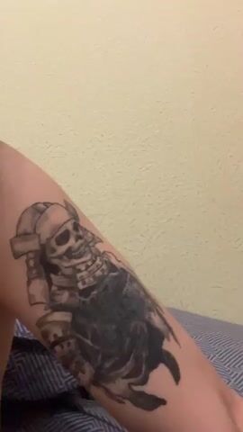 The video shows a person's arm with tattoos. The first tattoo appears to be a skeleton in samurai armor, with a black cloak. The second tattoo is of a bee. The person is wearing a blue and white striped shirt. The background consists of a light-colored wall and a wooden surface.